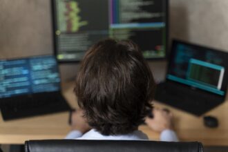 widok głowy od tyłu – człowiek siedzący przed monitorami trzech komputerów